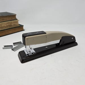 May include: A vintage black and beige metal stapler with the word "Swingline" printed on the top. The stapler is sitting on a white surface with several staples scattered around it.