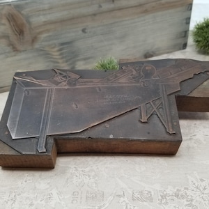 May include: A vintage wood printing block with a metal design of a small train engine. The block is made of wood and metal and has a brown color. The text on the block reads "Milwaukee Road,  Highway 421,  Chicago, Ill."