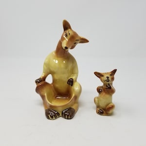 May include: A set of two ceramic figurines of a kangaroo and a joey. The kangaroo is sitting down with its tail curled around its legs. The joey is standing up and looking at the kangaroo. Both figurines are painted in shades of brown and yellow.