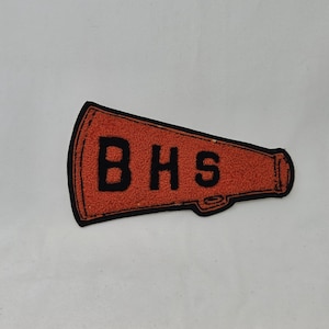 May include: A megaphone-shaped patch featuring a textured orange surface with a black border and the black letters BHS printed across the center. The design includes a small handle detail on the lower edge, rendered in a flat, embroidered style.