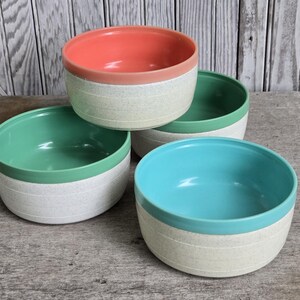 May include: A set of four round ceramic bowls with a speckled off-white base and horizontal grooved lines. Each bowl features a distinct solid-colored interior rim in coral, light green, dark green, and bright turquoise, creating a colorful display.