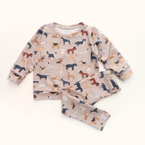 Könnte beinhalten: Beiges Langarm-Oberteil und passende Hose. Das Set ist mit einem sich wiederholenden Muster verschiedener Hunderassen in Braun, Schwarz und Weiß bedeckt. Das Outfit ist aus weichem Material und für Kinder geeignet.