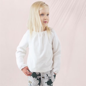May include: A young child wearing a white long-sleeved top with textured sleeves and a gathered neckline. The child is also wearing dark green pants with a white floral pattern. The background is a soft pink.