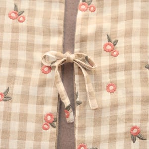 May include: Close-up of a beige and white checked fabric with pink floral embroidery. A matching bow is tied in the centre. The fabric appears to be a light, breathable material.