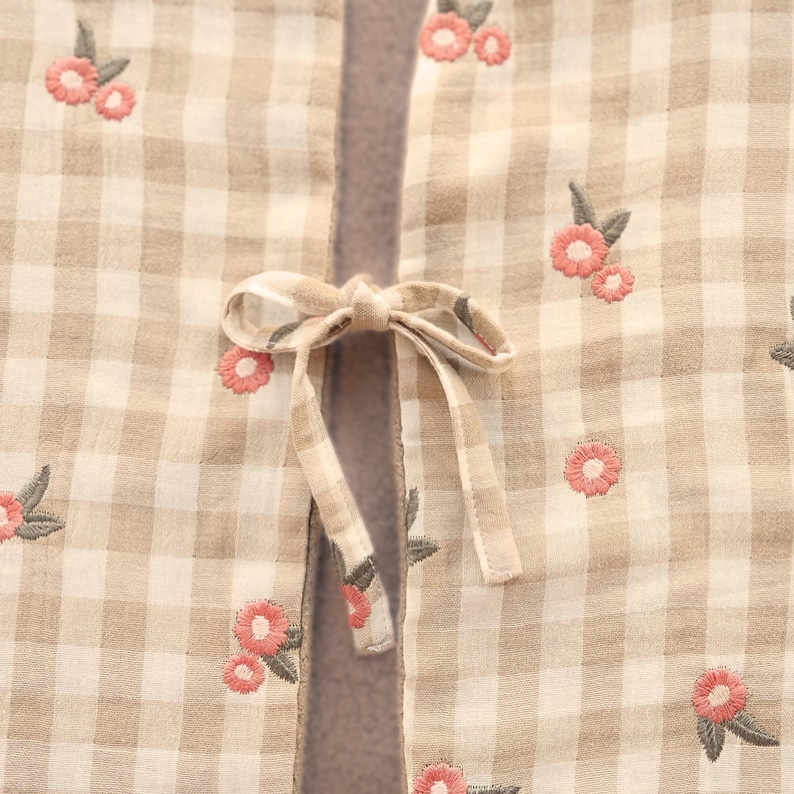 May include: Close-up of a beige and white checked fabric with pink floral embroidery. A matching bow is tied in the centre. The fabric appears to be a light, breathable material.