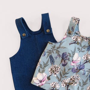 May include: Two children's denim overalls and a floral print overall. The denim overall is a dark blue color. The floral print overall has a light blue background with white, purple, and pink flowers. Both overalls have adjustable straps with gold-colored buttons.