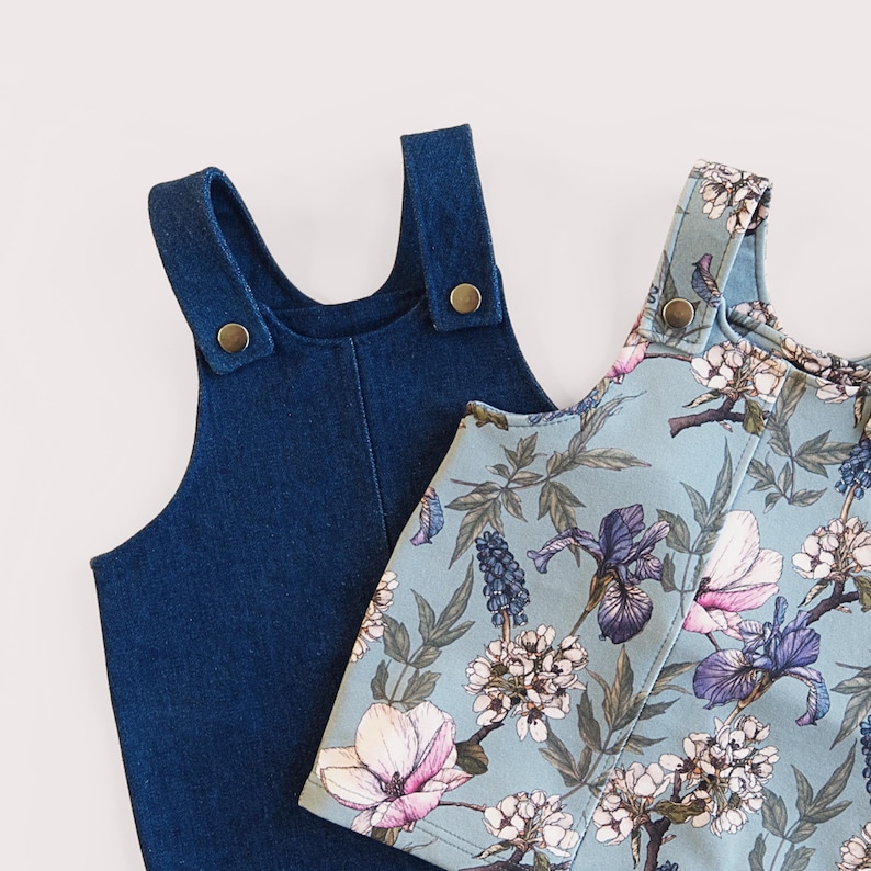 May include: Two children's denim overalls and a floral print overall. The denim overall is a dark blue color. The floral print overall has a light blue background with white, purple, and pink flowers. Both overalls have adjustable straps with gold-colored buttons.