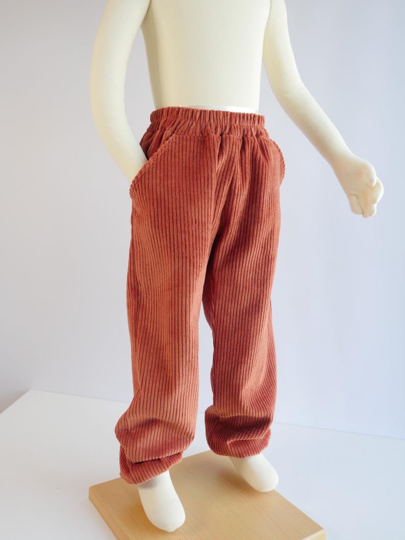 May include: Rust-coloured corduroy trousers featuring a comfortable elasticated waistband and two side pockets. The trousers are crafted from a textured, vertically ribbed fabric and have a relaxed, straight-leg fit that falls to the ankles.