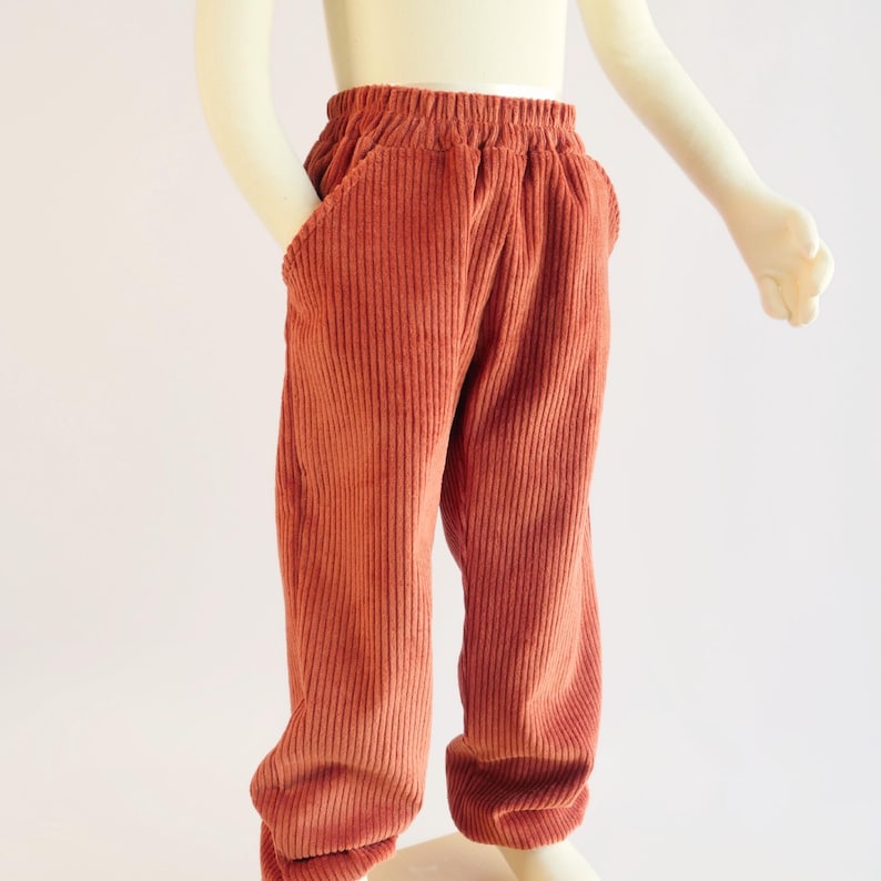 K&ouml;nnte beinhalten: Rostfarbene Cordhose mit elastischem Bund und Seitentaschen. Die Hose wird auf einer Schaufensterpuppe pr&auml;sentiert, die den l&auml;ssigen Stil und die Textur des Stoffes zeigt. Die Hose hat einen warmen, erdigen Farbton.