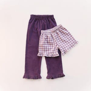May include: A pair of solid dark purple pants with an elastic waistband and ruffled hems, paired with a pair of light purple and white gingham patterned shorts featuring an elastic waistband and ruffled leg openings.