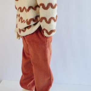 May include: A cream-coloured sherpa fleece pullover featuring a brown wavy line pattern paired with rust-coloured corduroy trousers. The pullover has long sleeves and a textured, fuzzy surface, while the trousers feature vertical ribbed detailing.
