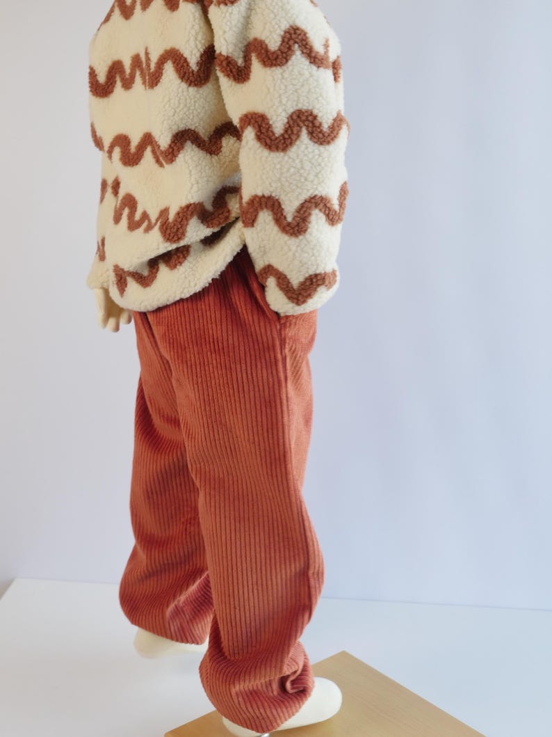 May include: A cream-coloured sherpa fleece pullover featuring a brown wavy line pattern paired with rust-coloured corduroy trousers. The pullover has long sleeves and a textured, fuzzy surface, while the trousers feature vertical ribbed detailing.