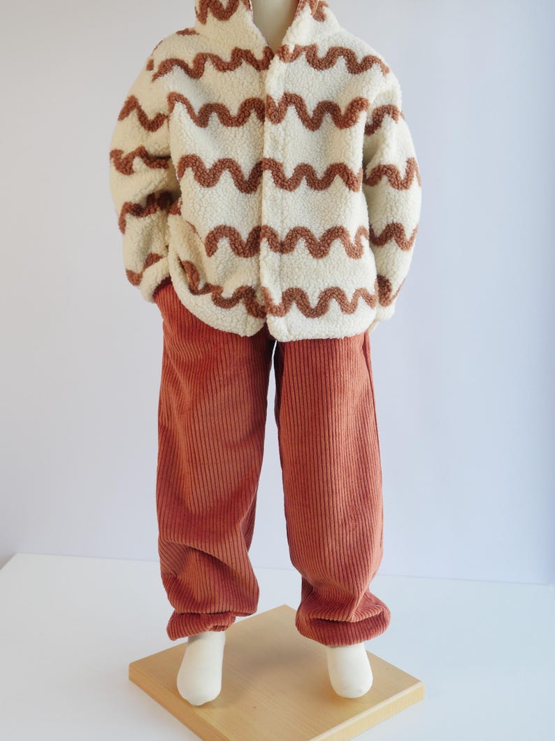 May include: A cream-coloured sherpa fleece jacket featuring a brown wavy line pattern and a front zip closure, paired with solid rust-coloured corduroy trousers that have a straight-leg fit and a textured ribbed surface throughout.