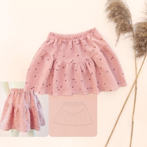 May include: A dusty pink skirt made of textured muslin fabric featuring a small dark heart pattern. The skirt has an elastic waistband and a tiered design with a gathered lower section. A line drawing of the skirt's construction is included.