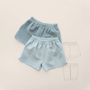 Puede incluir: Dos pares de pantalones cortos azul claro. Uno tiene un tejido de gofre texturizado y el otro es de lino liso. Los pantalones cortos tienen cinturillas elásticas. Un dibujo lineal de los pantalones cortos está en el fondo.