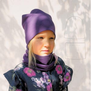 Puede incluir: Un niño con un gorro morado y un calentador de cuello a juego. El gorro tiene un diseño plegado único. El niño también lleva una chaqueta azul oscuro con un estampado floral. Un diagrama del gorro y el calentador de cuello está en el fondo.