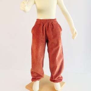K&ouml;nnte beinhalten: Rostfarbene Cordhose mit elastischem Bund und zwei Vordertaschen. Die Hose wird auf einer Schaufensterpuppe pr&auml;sentiert. Der Stoff hat eine vertikale Rippenstruktur. Die Hose ist f&uuml;r Kinder konzipiert.