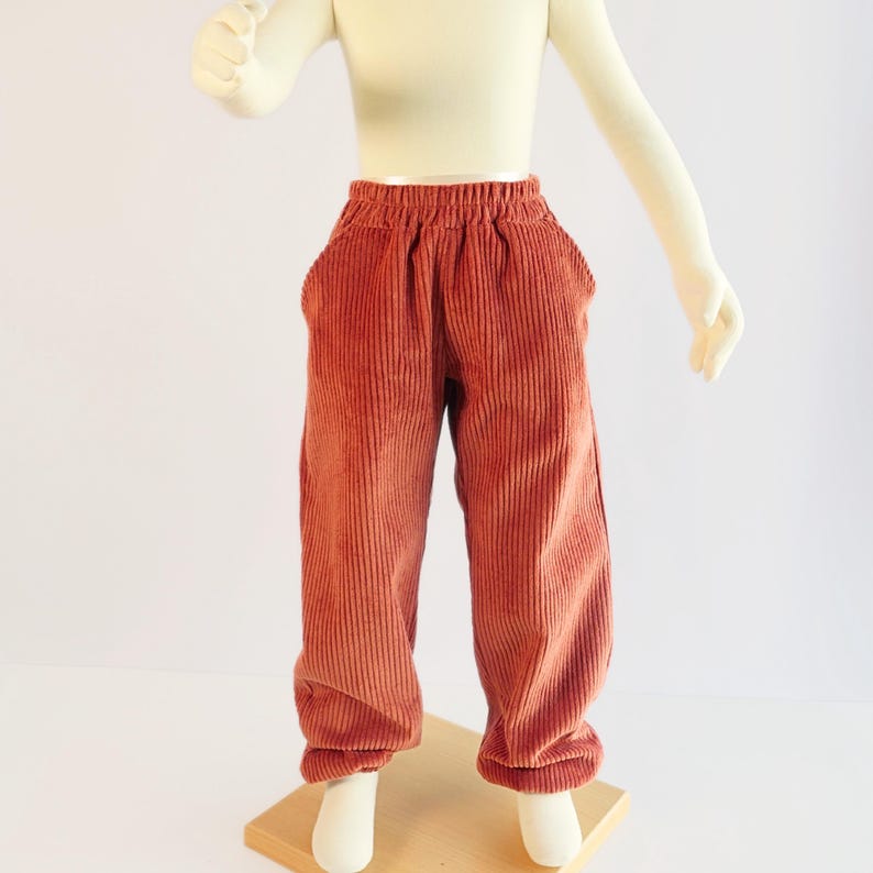 K&ouml;nnte beinhalten: Rostfarbene Cordhose mit elastischem Bund und zwei Vordertaschen. Die Hose wird auf einer Schaufensterpuppe pr&auml;sentiert. Der Stoff hat eine vertikale Rippenstruktur. Die Hose ist f&uuml;r Kinder konzipiert.