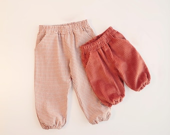 May include: Two pairs of pants with elastic waistbands and gathered cuffs. One pair features a light brown and white gingham check pattern, while the other pair is made of solid rust-colored corduroy fabric with vertical ridges.