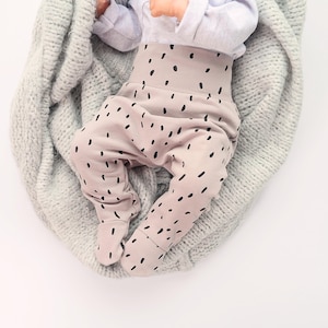 May include: Baby pants in a light tan color with a high waistband and black dot pattern. The pants have attached feet and are shown on a baby wrapped in a gray blanket. The baby is wearing a light gray long-sleeve shirt.