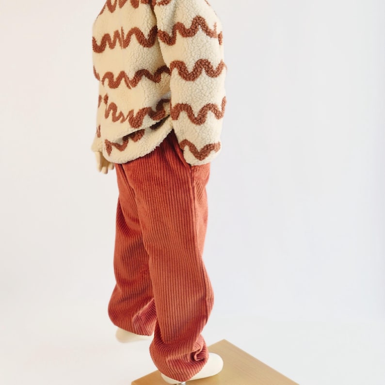 K&ouml;nnte beinhalten: Ein cremefarbene, flauschige Jacke mit braunen Wellenmustern, kombiniert mit rostfarbenen Cordhosen. Das Outfit wird auf einer Schaufensterpuppe pr&auml;sentiert, um die Textur und den Stil der Kinderkleidung zu zeigen.