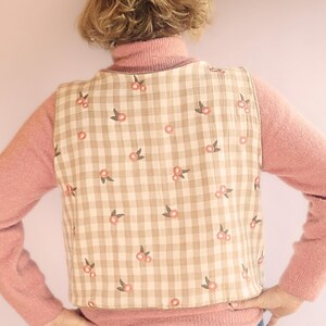 May include: A beige and white checked vest featuring a floral design of pink flowers and green leaves. The vest is worn over a pink turtleneck sweater.