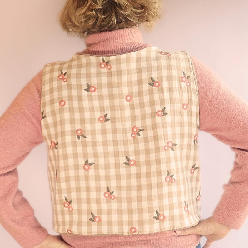 May include: A beige and white checked vest featuring a floral design of pink flowers and green leaves. The vest is worn over a pink turtleneck sweater.
