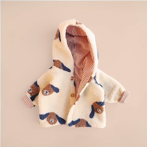 May include: A cream-coloured, hooded jacket for children. The jacket features a repeating pattern of brown and blue dog faces. The interior lining and cuffs are a red and brown gingham pattern. The jacket has two gold-coloured buttons.