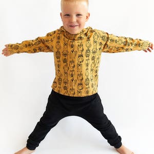 May include: A young boy wearing a mustard yellow long-sleeved shirt with a black and white owl print. The shirt has a hood. The boy is also wearing black pants.