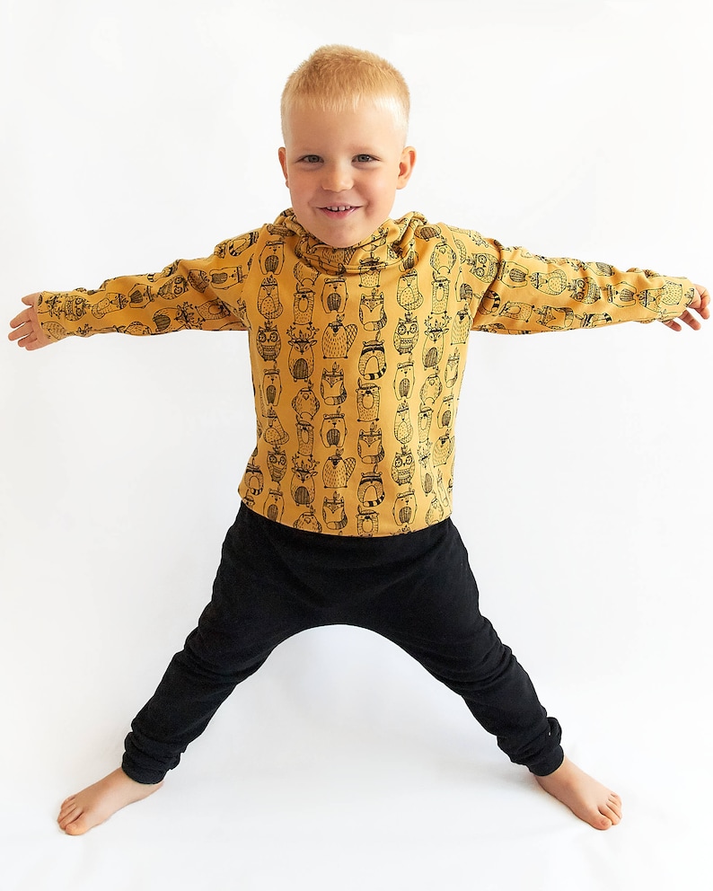 May include: A young boy wearing a mustard yellow long-sleeved shirt with a black and white owl print. The shirt has a hood. The boy is also wearing black pants.