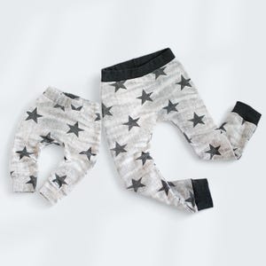 Op de afbeelding: Twee paar grijze leggings met een zwart sterrenpatroon. Het kleinere paar is gevouwen en het grotere paar ligt uitgespreid.