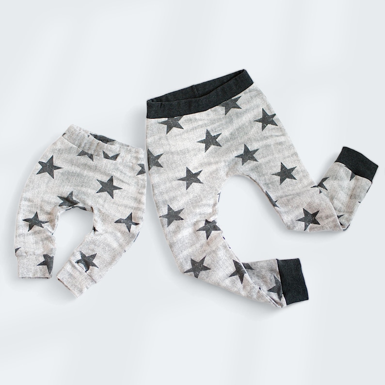 Op de afbeelding: Twee paar grijze leggings met een zwart sterrenpatroon. Het kleinere paar is gevouwen en het grotere paar ligt uitgespreid.