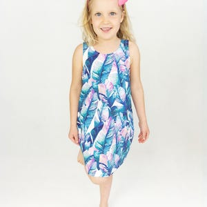 May include: A sleeveless dress featuring a vibrant tropical leaf pattern in shades of blue, purple, and pink. The dress is worn by a young child with blonde hair and a pink bow. The dress has a scoop neckline and a relaxed fit.