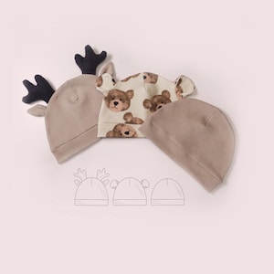 May include: Three baby hats in neutral tones. One hat has black antlers, another has bear ears, and the third is plain. The bear-themed hat features a repeating pattern of teddy bear faces. A line drawing of the hats is at the bottom.