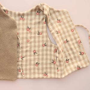 May include: A beige and white checked vest with embroidered pink floral accents. The vest has a soft, plush interior and a tie closure. The floral pattern is repeated across the checked fabric.