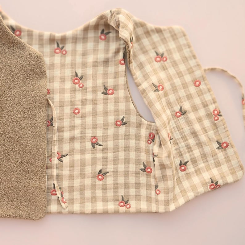 May include: A beige and white checked vest with embroidered pink floral accents. The vest has a soft, plush interior and a tie closure. The floral pattern is repeated across the checked fabric.