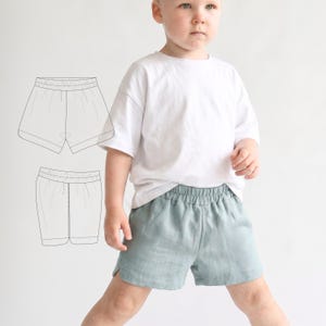 May include: A young child wearing a white t-shirt and light blue shorts. The shorts have an elastic waistband and side slits. A line drawing of the shorts is visible on the left side of the image. The shorts are made of a soft, breathable fabric.