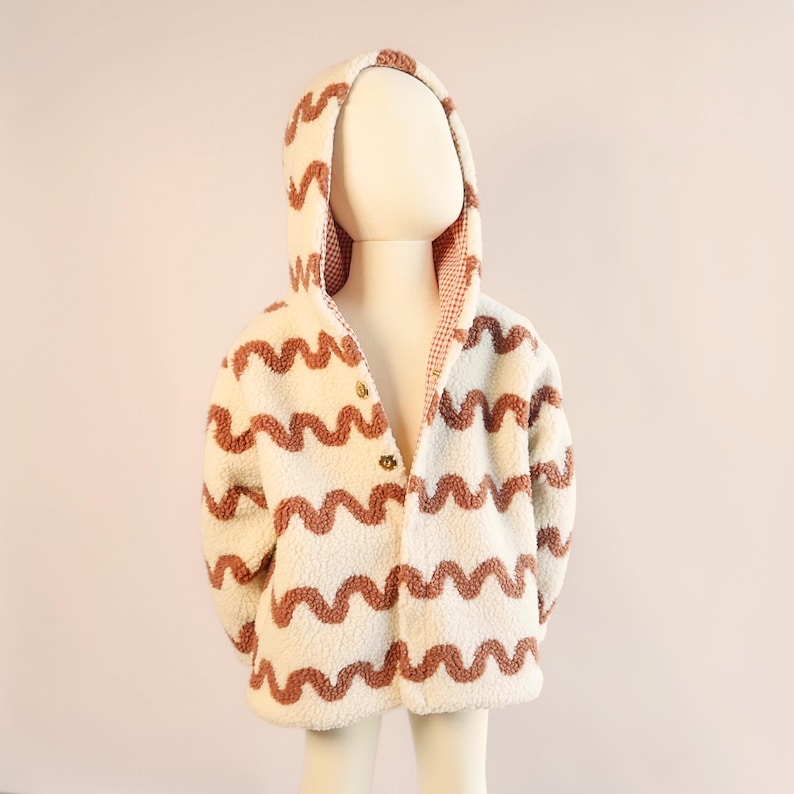 Puede incluir: Chaqueta con capucha de color crema con un patr&oacute;n ondulado marr&oacute;n. La chaqueta tiene cierre de botones y una capucha forrada con un patr&oacute;n de cuadros rojos y blancos. La chaqueta se muestra en un maniqu&iacute;.
