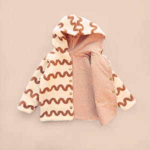 Può includere: Giacca con cappuccio color crema con linee ondulate marroni e bottoni dorati. La fodera interna è a quadri rossi e bianchi. La giacca è realizzata in un materiale morbido e strutturato.