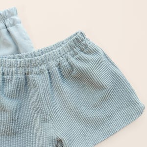Puede incluir: Primer plano de unos pantalones cortos de pana azul claro con cintura el&aacute;stica. Los pantalones cortos tienen un aspecto texturizado y acanalado y un ajuste relajado. La tela es de color azul claro liso, y los pantalones cortos parecen suaves y c&oacute;modos. La prenda es probablemente para uso casual.