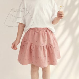 Könnte beinhalten: Hellrosa Rock mit Herzmuster. Der Rock hat ein gestuftes Design und einen elastischen Bund. Ein weißes T-Shirt wird mit dem Rock getragen. Eine Skizze des Rocks befindet sich in der oberen linken Ecke.