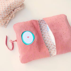 Può includere: Una custodia in tessuto rosa polvere con macchioline dorate e una fodera interna floreale. La custodia presenta un ritaglio circolare centrale che rivela un dispenser di salviette in plastica turchese con coperchio bianco e un cuore rosso.