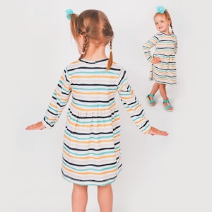 May include: A long-sleeved striped dress in shades of yellow, blue, and black. The dress features a round neckline and a gathered waist. A young girl is wearing the dress, with her arms outstretched. Another girl is wearing the same dress in the background.