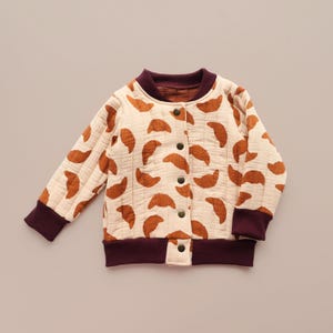 May include: Cream-colored quilted bomber jacket with a repeating pattern of brown croissant shapes. The jacket features a burgundy collar, cuffs, and hem, and fastens with small, dark buttons. A cozy and stylish outerwear option.