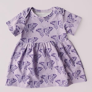 May include: A light purple baby dress with short sleeves and a gathered skirt. The dress is covered in a repeating pattern of dark purple butterfly illustrations. The neckline and sleeves have a simple, clean finish, and the dress appears to be made of soft fabric.