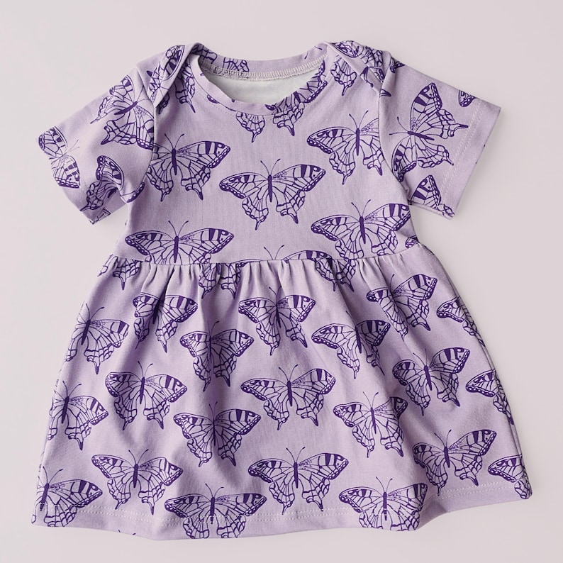 May include: A light purple baby dress with short sleeves and a gathered skirt. The dress is covered in a repeating pattern of dark purple butterfly illustrations. The neckline and sleeves have a simple, clean finish, and the dress appears to be made of soft fabric.