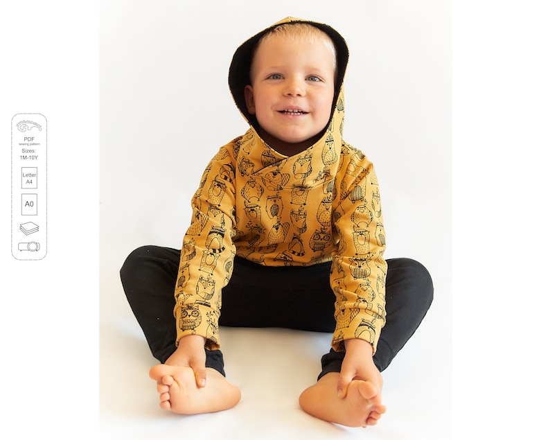 May include: A young child wearing a mustard yellow hoodie with a black and white owl print. The hoodie has a cowl neck and long sleeves. The child is sitting on a white surface with their legs crossed.