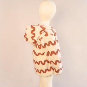 Puede incluir: Chaqueta con capucha color crema con un patr&oacute;n ondulado marr&oacute;n. La chaqueta est&aacute; hecha de un material suave y texturizado y est&aacute; dise&ntilde;ada para ni&ntilde;os. La capucha est&aacute; forrada con una tela a cuadros roja y blanca.