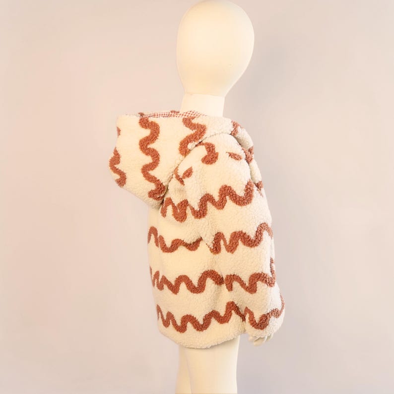 Puede incluir: Chaqueta con capucha color crema con un patr&oacute;n ondulado marr&oacute;n. La chaqueta est&aacute; hecha de un material suave y texturizado y est&aacute; dise&ntilde;ada para ni&ntilde;os. La capucha est&aacute; forrada con una tela a cuadros roja y blanca.