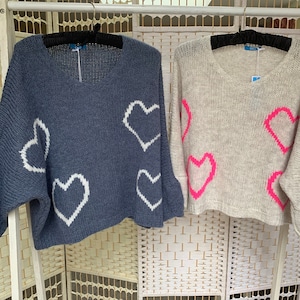 May include: Two V-neck jumpers, one in navy blue with white heart shapes, and another in light beige with bright pink hearts. Both jumpers have long sleeves and are hanging on black hangers.